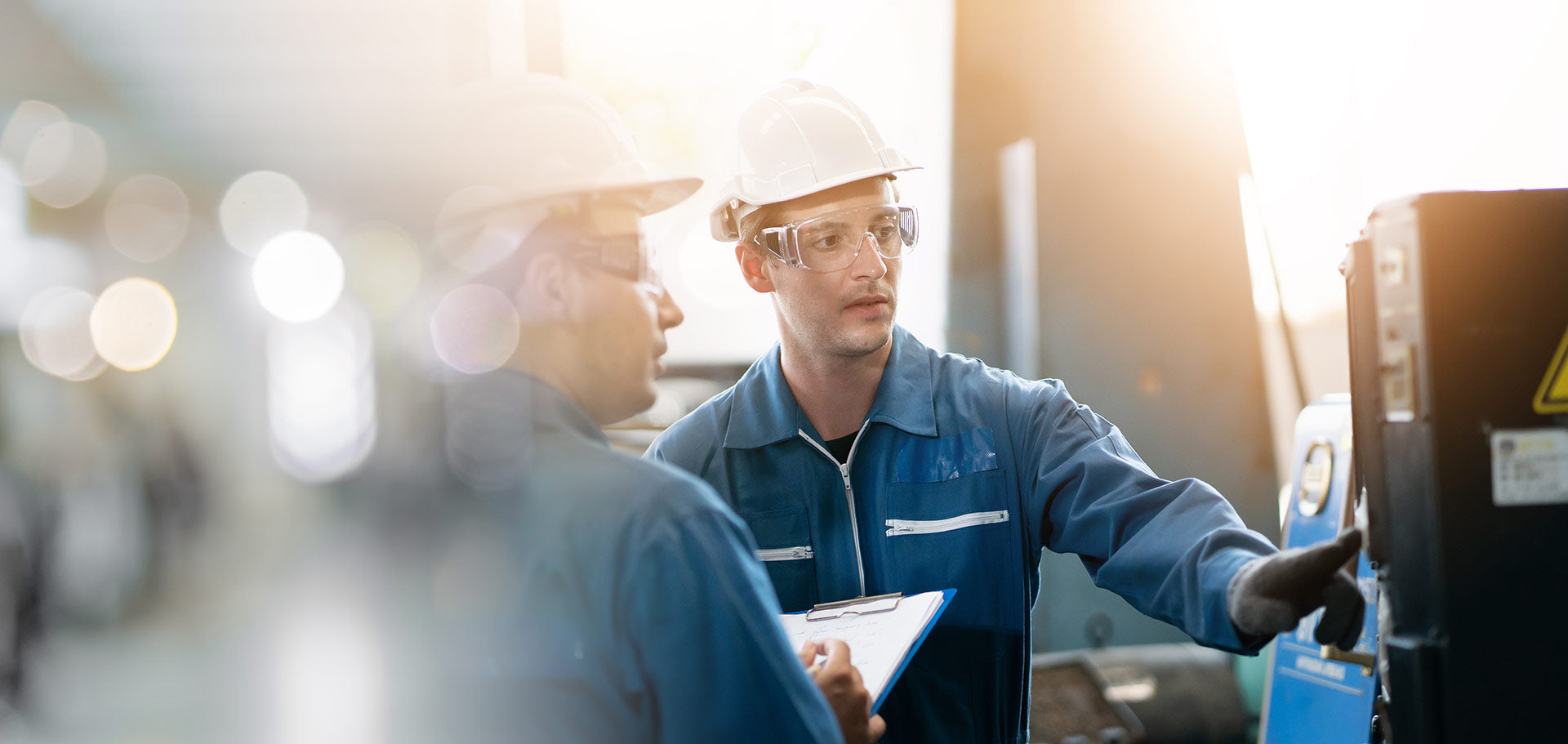 Two workers in a factory setting, one holding a clipboard and the other looking at machinery.