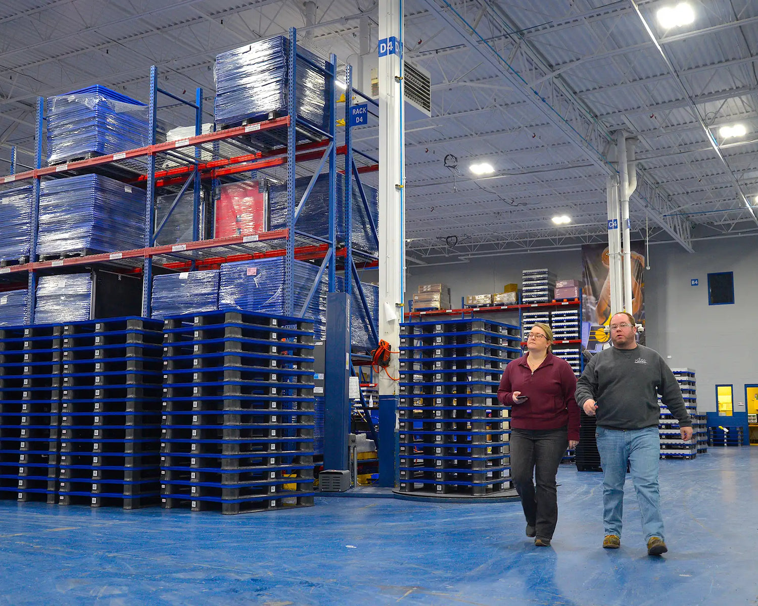 Two people walking through a warehouse with blue pallets and storage racks.